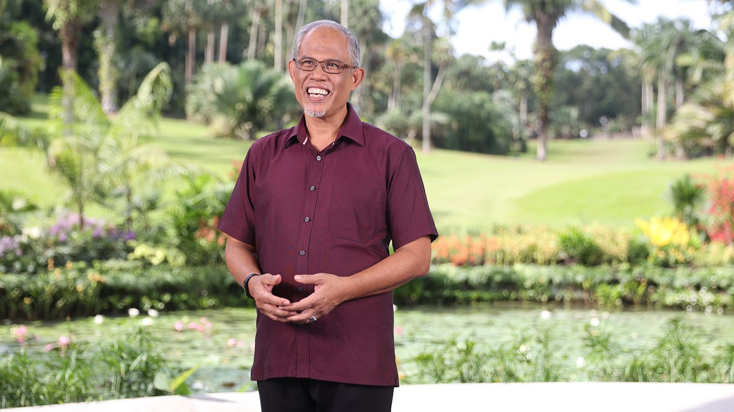 Smiling man in maroon shirt with hands clasped, standing in a tropical garden.